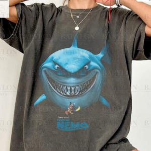 May include: A dark gray t-shirt featuring a large, blue cartoon shark with a menacing grin. The shirt has the text "NEMO" and the Disney Pixar logo. The words "Comfort Colors Pepper" are printed on the top right. The Babylon Prints logo is on the top left.