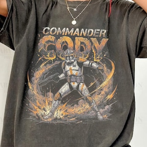 May include: Dark grey t-shirt featuring a graphic of Commander Cody from Star Wars. The design includes the text "Commander Cody" and an illustration of the character in action, with orange and yellow accents. The shirt is labeled "Comfort Colors Pepper".