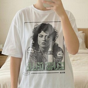 Könnte beinhalten: Weißes T-Shirt mit einem Schwarz-Weiß-Bild einer Frau, dem Text "Ellen Ripley" und "Alien" darunter. Das T-Shirt hat auch den Text "1979" aufgedruckt. Das T-Shirt ist von Babylon Prints.