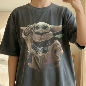 以下が含まれることがあります： マンダロリアンに登場するグローグー（ベイビーヨーダとしても知られる）のグラフィックがプリントされた濃いグレーのTシャツ。Tシャツは半袖でクルーネックです。「Comfort Colors Pepper」の文字が見えます。モデルはXLサイズを着用しています。