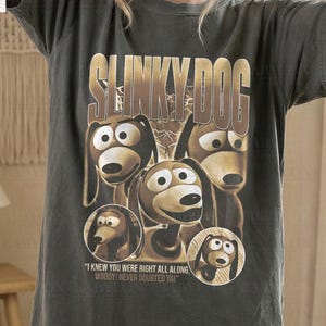 May include: Dark grey t-shirt featuring a graphic of Slinky Dog from Toy Story. The design includes the text "SLINKY DOG" and several images of the character. The shirt also has the text "I KNEW YOU WERE RIGHT ALL ALONG. WOODY! NEVER DOUBTED YA!"