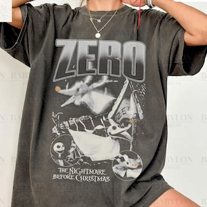 May include: Dark grey t-shirt featuring the word "ZERO" above a graphic from "The Nightmare Before Christmas." The design includes Zero the ghost dog and other characters from the movie. The shirt is a Comfort Colors Pepper tee.