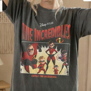 Può includere: Una maglietta grigia con una grafica de "Gli Incredibili" di Disney Pixar. Il design include il titolo del film e i personaggi in rosso, nero e giallo. La maglietta è etichettata "Comfort Colors Pepper" e "Original Artist of This Design."