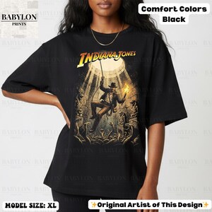 May include: Black t-shirt featuring a graphic of Indiana Jones in a tomb, holding a torch and rope. The text "INDIANA JONES" is in yellow. The shirt is a comfort color and the model is wearing size XL.