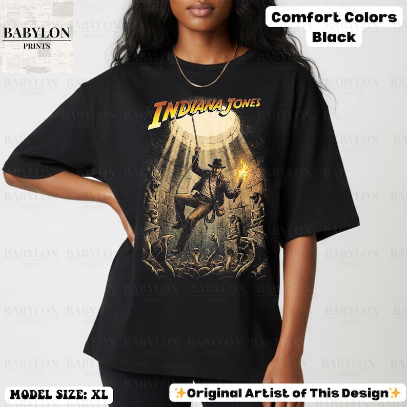 May include: Black t-shirt featuring a graphic of Indiana Jones in a tomb, holding a torch and rope. The text "INDIANA JONES" is in yellow. The shirt is a comfort color and the model is wearing size XL.