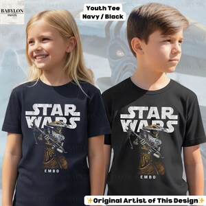 Puede incluir: Dos camisetas juveniles, azul marino y negra, cada una con un gr&aacute;fico de Star Wars. El dise&ntilde;o incluye las palabras "STAR WARS" en blanco sobre una ilustraci&oacute;n de un personaje con una ballesta. El nombre del personaje, "EMBO", est&aacute; impreso debajo.