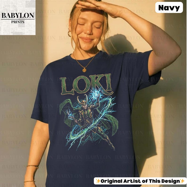 May include: Navy blue t-shirt featuring the word "LOKI" in green above a graphic of a character with green and gold attire, surrounded by blue energy. The shirt is from "BABYLON PRINTS" and has "Original Artist of This Design" text.