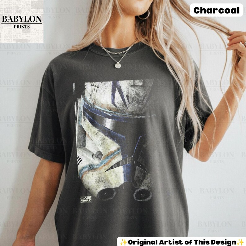 May include: A charcoal gray short-sleeve t-shirt featuring a large graphic print of a weathered white and blue clone trooper helmet. The helmet design includes dark blue accents and distressed markings. The text "CLONE WARS" is printed in small white letters.