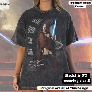 Może przedstawiać: Ciemnoszara koszulka z grafiką Obi-Wana Kenobiego trzymającego niebieski miecz świetlny. Koszulka ma napis "Obi-Wan Kenobi" w stylu odręcznym. Koszulka jest prana w kolorze pieprzu. Modelka ma 170 cm wzrostu i nosi rozmiar S.