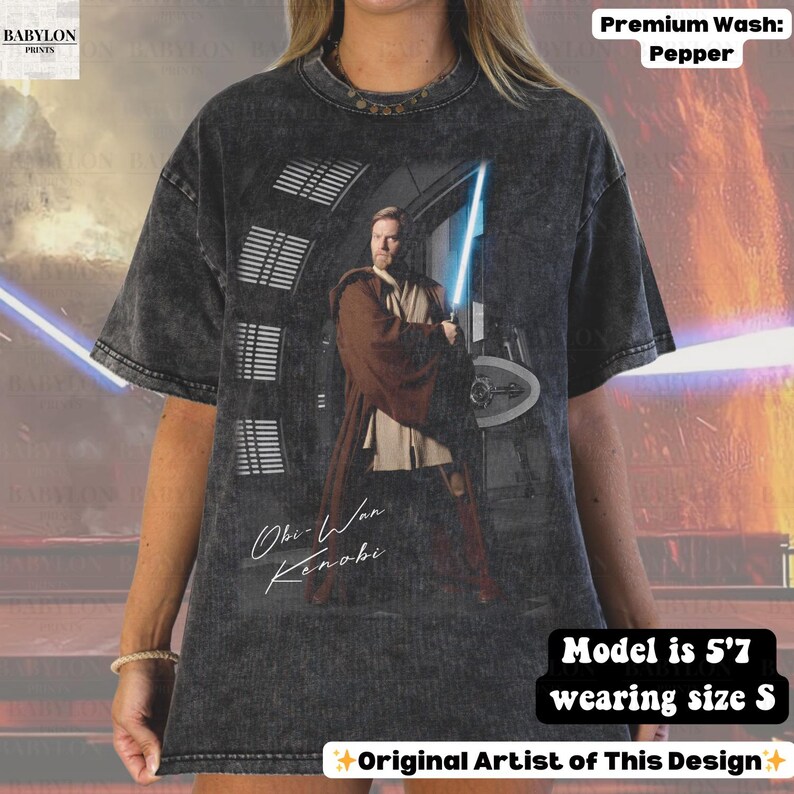 Może przedstawiać: Ciemnoszara koszulka z grafiką Obi-Wana Kenobiego trzymającego niebieski miecz świetlny. Koszulka ma napis "Obi-Wan Kenobi" w stylu odręcznym. Koszulka jest prana w kolorze pieprzu. Modelka ma 170 cm wzrostu i nosi rozmiar S.