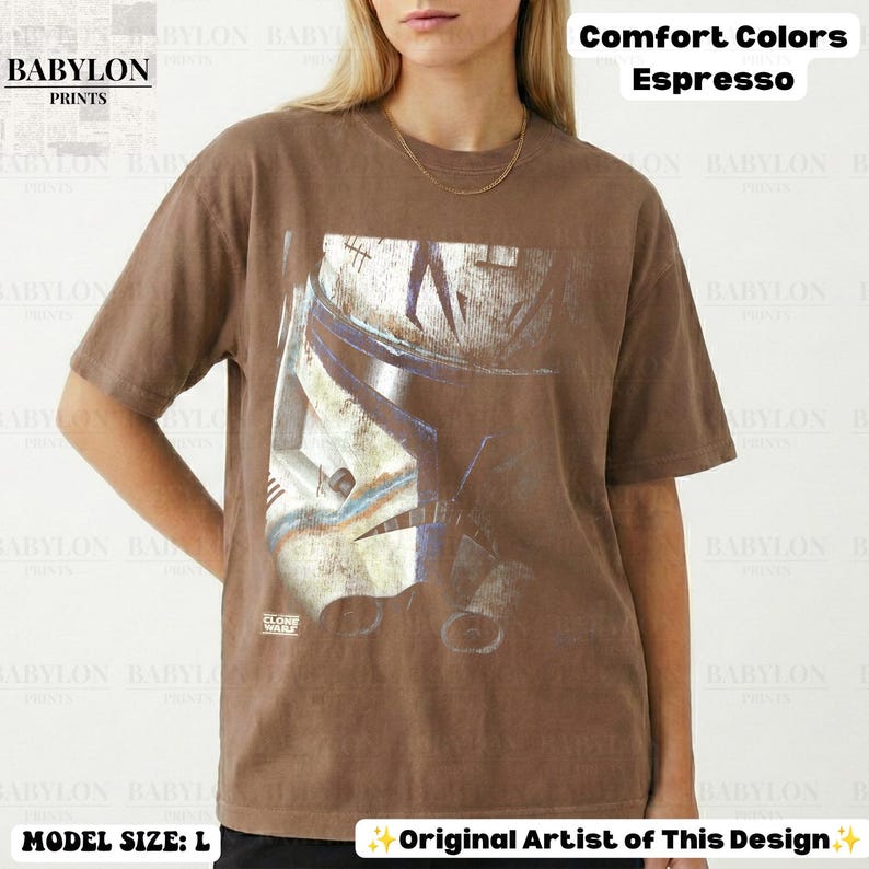 May include: A brown short-sleeve t-shirt featuring a large graphic print of a white and blue clone trooper helmet. The shirt includes the text Comfort Colors Espresso, Babylon Prints, and Clone Wars. The garment is displayed in a size large.