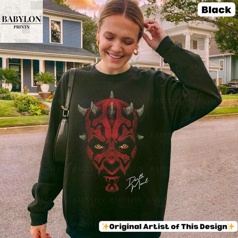 Może przedstawiać: Czarna bluza z okrągłym dekoltem z czerwono-czarną grafiką twarzy Darth Maula. Projekt zawiera rogi i ż&oacute;łte oczy, z napisem "Darth Maul" w białym skrypcie. Bluza ma napis "BABYLON PRINTS" w lewym g&oacute;rnym rogu i "Original Artist of This Design" na dole.