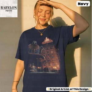 May include: Navy blue t-shirt with a graphic of a building and fountain, and the text "GUSTEAU'S". The shirt also has the text "Original Artist of This Design". The t-shirt is a crew neck.