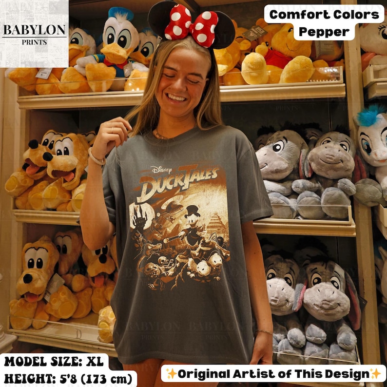 May include: A gray t-shirt featuring a Disney DuckTales graphic. The shirt is worn by a person in front of a shelf of plush toys. The text "Comfort Colors Pepper" is visible. The model's size is XL and height is 5'8 (173 cm).