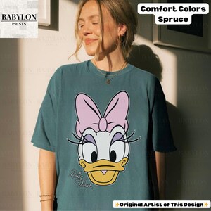 May include: Spruce-colored t-shirt featuring a Daisy Duck graphic. The design includes a pink bow, white face, and yellow beak. The text "Daisy Duck" is written in cursive. The shirt is from "Comfort Colors" and "Original Artist of This Design".