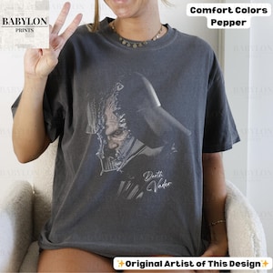 K&ouml;nnte beinhalten: Ein dunkelgraues T-Shirt mit einer Grafik von Darth Vader. Der Text "Darth Vader" ist in kursiver Schrift unter der Grafik geschrieben. Das Shirt ist mit "Comfort Colors Pepper" und "Original Artist of This Design" gekennzeichnet.