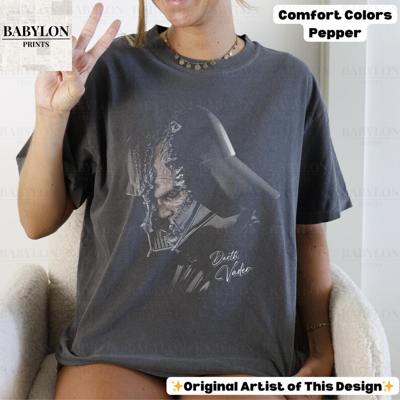 K&ouml;nnte beinhalten: Ein dunkelgraues T-Shirt mit einer Grafik von Darth Vader. Der Text "Darth Vader" ist in kursiver Schrift unter der Grafik geschrieben. Das Shirt ist mit "Comfort Colors Pepper" und "Original Artist of This Design" gekennzeichnet.