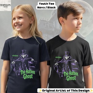 Puede incluir: Una camiseta juvenil de manga corta en azul marino y negro, con un estampado gr&aacute;fico de un personaje con sombrero de copa que sostiene una sombrerera. El texto dice Haunted Mansion y The Hatbox Ghost en letras verdes.