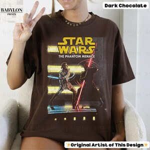 Puede incluir: Camiseta marr&oacute;n chocolate oscuro con un dise&ntilde;o de Star Wars. El dise&ntilde;o incluye el texto "STAR WARS THE PHANTOM MENACE" e ilustraciones de personajes con sables de luz. La camiseta tiene cuello redondo y mangas cortas.