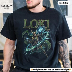 May include: Black t-shirt featuring a green and gold illustration of Loki with the word "LOKI" above. The design includes green lightning and a green cape. The shirt has the text "Original Artist of This Design" at the bottom.