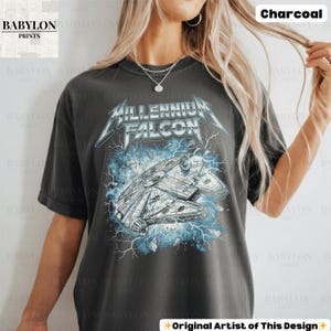 May include: Charcoal-colored t-shirt featuring a graphic of the Millennium Falcon with the text "Millennium Falcon" in a stylized font. The design includes blue lightning and a distressed effect, creating a vintage look. The shirt is from Babylon Prints.