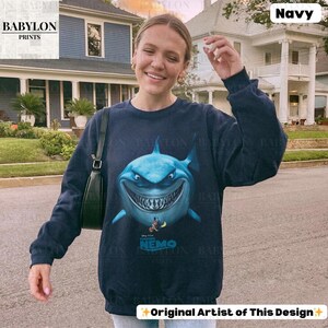 May include: Navy blue sweatshirt featuring a large, cartoonish shark with a menacing grin. The shark is light blue with white teeth and a dark blue outline. Below the shark is the text "Finding Nemo". The sweatshirt has long sleeves and a crew neck.