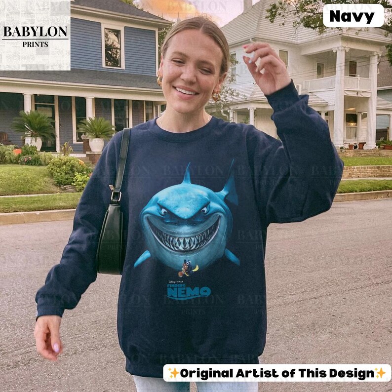 May include: Navy blue sweatshirt featuring a large, cartoonish shark with a menacing grin. The shark is light blue with white teeth and a dark blue outline. Below the shark is the text "Finding Nemo". The sweatshirt has long sleeves and a crew neck.