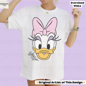 Puede incluir: Camiseta blanca de gran tama&ntilde;o con un dise&ntilde;o de Daisy Duck. El personaje de dibujos animados tiene un lazo rosa, pesta&ntilde;as negras y un pico amarillo. El texto "Daisy Duck" est&aacute; debajo. La camiseta est&aacute; etiquetada como "Oversized White".