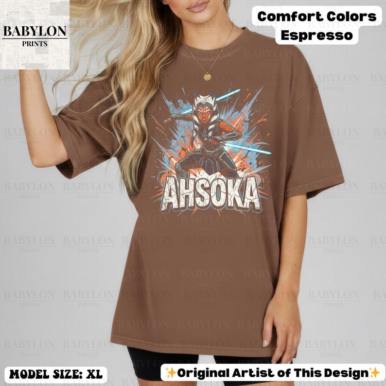 Może przedstawiać: Brązowa koszulka w kolorze espresso z grafiką Ahsoki Tano z mieczami świetlnymi i napisem "AHSOKA". Koszulka marki Comfort Colors, modelka nosi rozmiar XL.