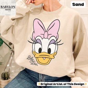 May include: Sand-colored sweatshirt featuring a large graphic of Daisy Duck with a pink bow. The design includes the text "Daisy Duck" in cursive. The sweatshirt has long sleeves and a crew neck. The text "Original Artist of This Design" is also visible.