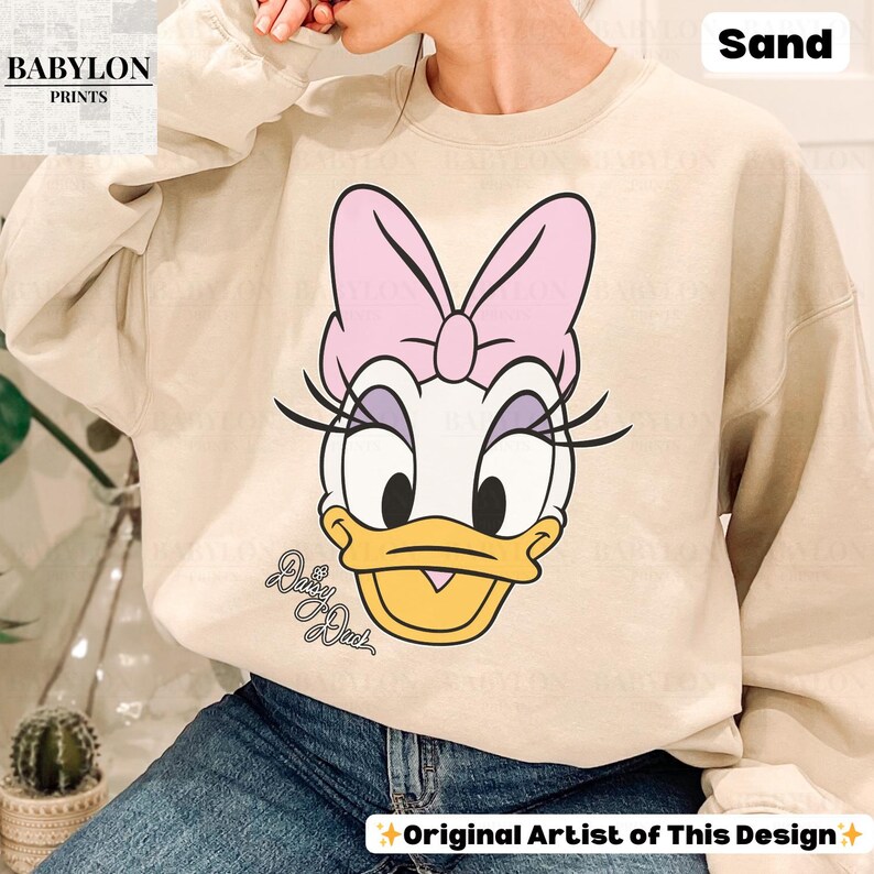 May include: Sand-colored sweatshirt featuring a large graphic of Daisy Duck with a pink bow. The design includes the text "Daisy Duck" in cursive. The sweatshirt has long sleeves and a crew neck. The text "Original Artist of This Design" is also visible.