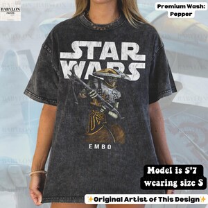 Puede incluir: Camiseta gris oscuro de Star Wars con el texto blanco "STAR WARS". La camiseta presenta un gr&aacute;fico del personaje Embo sosteniendo un arma, con el texto "EMBO" debajo. La modelo mide 1,70 m y usa la talla S. La camiseta es un lavado premium pimienta.