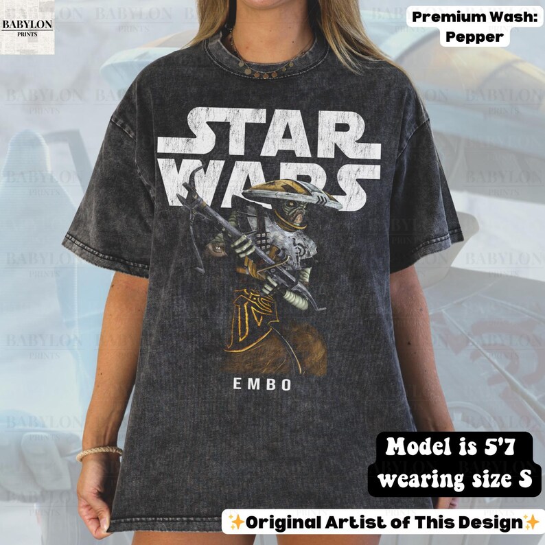 Puede incluir: Camiseta gris oscuro de Star Wars con el texto blanco "STAR WARS". La camiseta presenta un gr&aacute;fico del personaje Embo sosteniendo un arma, con el texto "EMBO" debajo. La modelo mide 1,70 m y usa la talla S. La camiseta es un lavado premium pimienta.