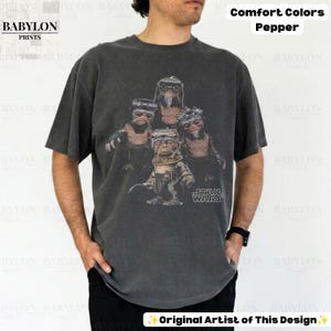 Op de afbeelding: Een antracietgrijs t-shirt met korte mouwen en een grafische print van vier Ewok-personages uit Star Wars. Het shirt is gelabeld als Comfort Colors Pepper en bevat het Star Wars-logo in het wit rechtsonder op de afbeelding.