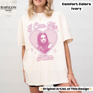 May include: Ivory-colored t-shirt with the text "I Can Fix Him" in pink, featuring a heart-shaped graphic with a portrait. The shirt is from Comfort Colors and has the Babylon Prints logo.