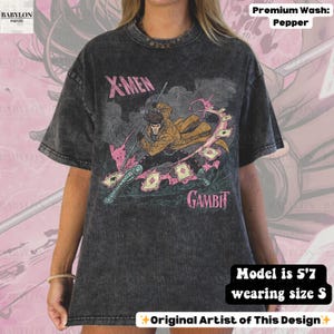 May include: Dark grey t-shirt featuring a graphic of Gambit from X-Men. The design includes the text "X-MEN" and "GAMBIT" in pink and white. The shirt is a premium wash in pepper color. The model is 5'7 and wearing a size S.