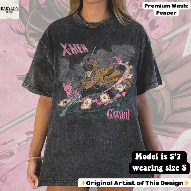 May include: Dark grey t-shirt featuring a graphic of Gambit from X-Men. The design includes the text "X-MEN" and "GAMBIT" in pink and white. The shirt is a premium wash in pepper color. The model is 5'7 and wearing a size S.