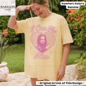 May include: A light yellow t-shirt with the text "I Can Fix Him" in pink, featuring a heart-shaped graphic with a portrait. The shirt also has the text "Comfort Colors Banana" and "Original Artist of This Design".