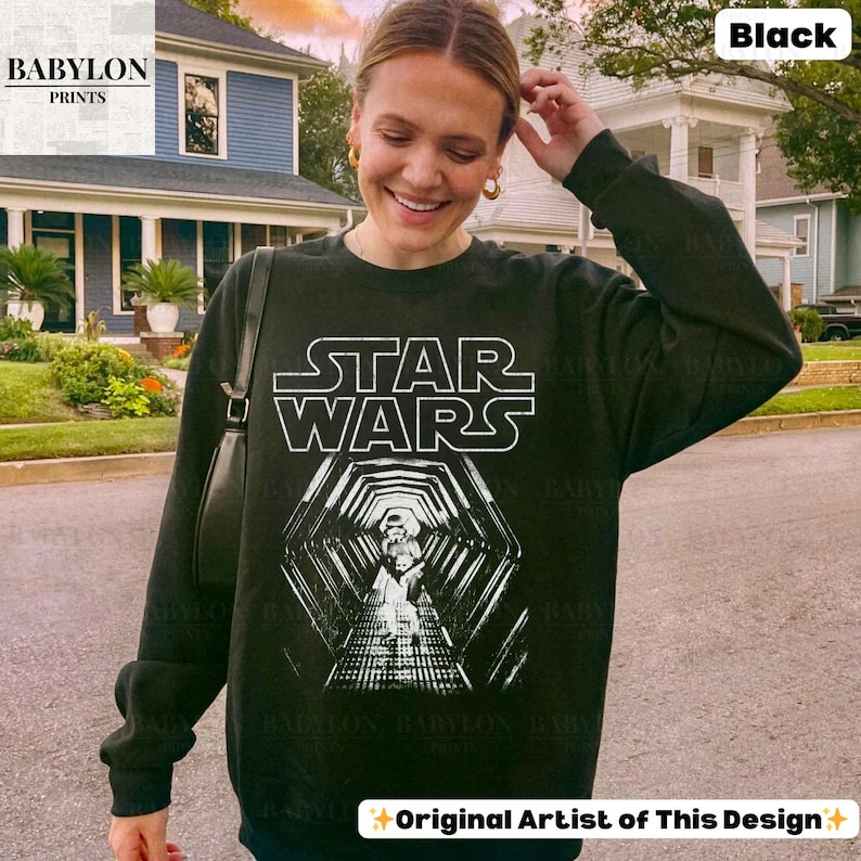 May include: Black Star Wars sweatshirt with a white graphic of the Star Wars logo and a tunnel design. The sweatshirt has long sleeves and a crew neck. The text "BABYLON PRINTS" is in the upper left corner, and "Original Artist of This Design" is at the bottom.
