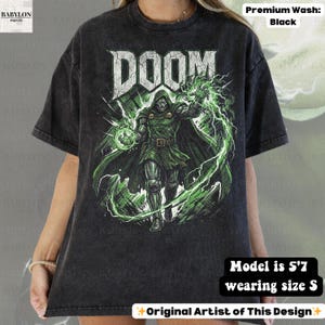 May include: Black t-shirt with a distressed graphic of Doctor Doom and the word "DOOM" in white. The design features green lightning and a green-clad figure. The shirt is labeled "Premium Wash: Black" and "Original Artist of This Design."