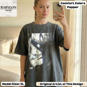 May include: A charcoal gray short-sleeve t-shirt featuring a large graphic print of a white and blue clone trooper helmet. The shirt has a crew neckline and a relaxed fit. The text "Comfort Colors Pepper" and "Model Size: XL" are visible.
