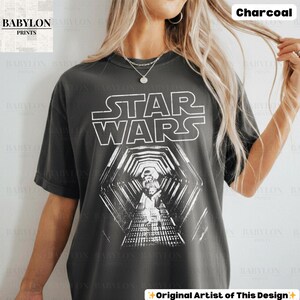 May include: Charcoal-colored Star Wars graphic t-shirt. The shirt features the Star Wars logo in white block letters above a detailed black and white image of a hallway with a Stormtrooper. The text "Original Artist of This Design" is at the bottom.