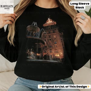 May include: Black long-sleeve top with a detailed illustration of a fountain and a building with the text "GUSTEAU'S" in red. The top also has the text "Long Sleeve Black" and "Original Artist of This Design".