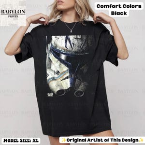 May include: A black short-sleeve t-shirt featuring a large graphic print of a weathered white and blue clone trooper helmet. The design includes the text Clone Wars in the bottom left corner of the graphic. The shirt is a Comfort Colors brand.