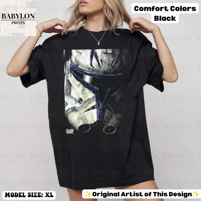 May include: A black short-sleeve t-shirt featuring a large graphic print of a weathered white and blue clone trooper helmet. The design includes the text Clone Wars in the bottom left corner of the graphic. The shirt is a Comfort Colors brand.