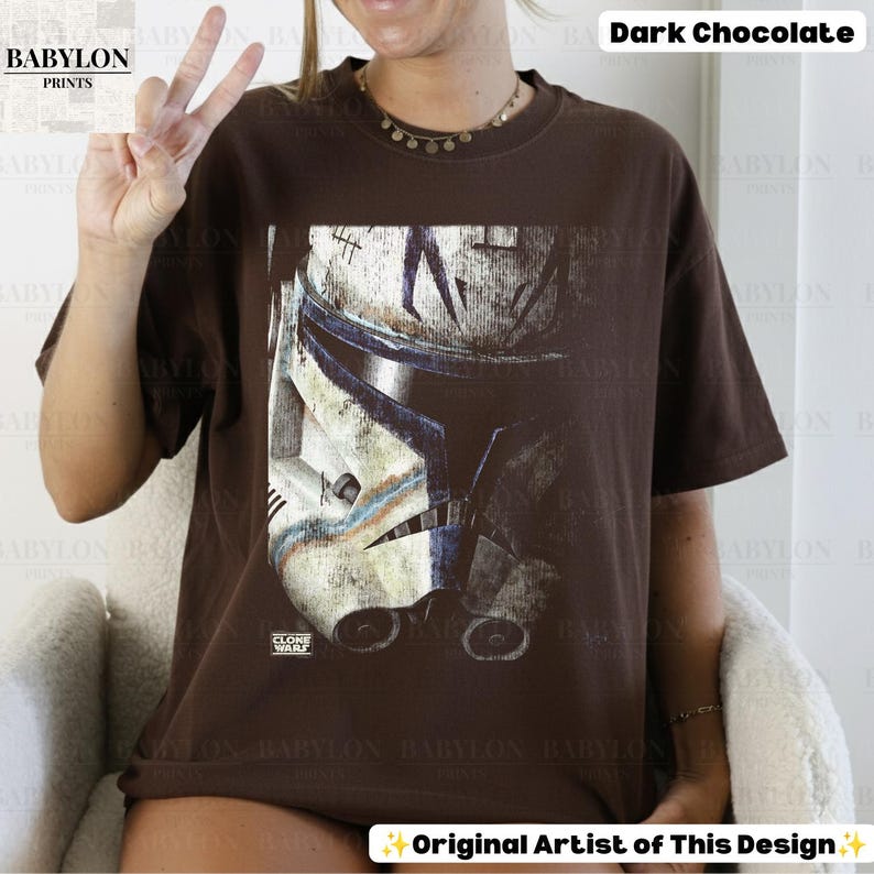 May include: A dark chocolate brown short-sleeve t-shirt featuring a large graphic print of a weathered white and blue clone trooper helmet. The design includes the text "CLONE WARS" in small white letters near the bottom of the helmet illustration.