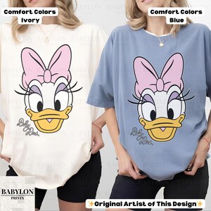 May include: Ivory and blue Comfort Colors t-shirts featuring a Daisy Duck graphic. The cartoon character has a pink bow, long eyelashes, and a yellow beak. The text "Daisy Duck" is below the character's face. The Babylon Prints logo is in the lower left corner.