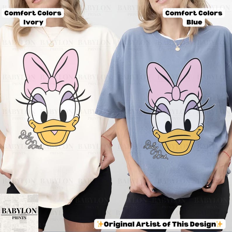 May include: Ivory and blue Comfort Colors t-shirts featuring a Daisy Duck graphic. The cartoon character has a pink bow, long eyelashes, and a yellow beak. The text "Daisy Duck" is below the character's face. The Babylon Prints logo is in the lower left corner.