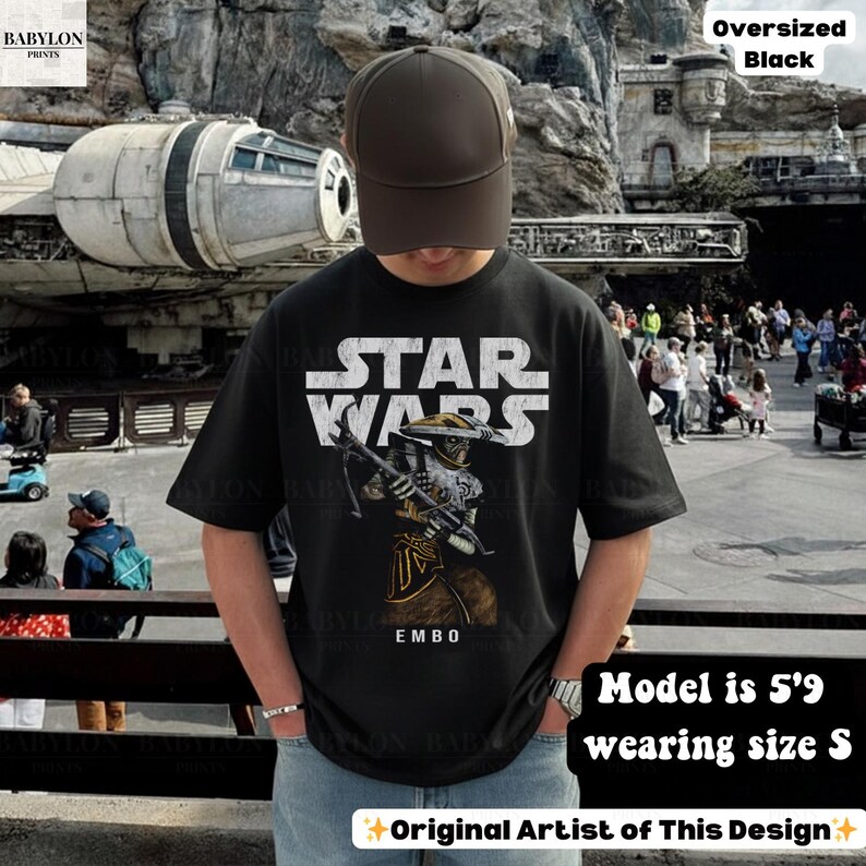 Puede incluir: Camiseta negra de gran tama&ntilde;o de Star Wars con un gr&aacute;fico de un personaje y el texto "EMBO". La camiseta tiene el texto "STAR WARS" en blanco. La modelo mide 1,75 m y lleva la talla S. El dise&ntilde;o es del artista original.