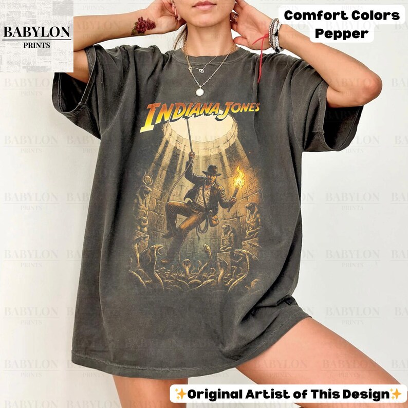May include: Dark gray t-shirt featuring the Indiana Jones movie poster. The graphic shows Indiana Jones holding a torch and descending into a temple. The text "INDIANA JONES" is in yellow. The shirt is labeled "Comfort Colors Pepper" and "Original Artist of This Design."